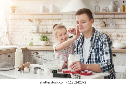 Happy Family Cooking And Having Fun Together In Kitchen. Cute Girl Smearing Her Dad Face With Flour, Smiling At Camera