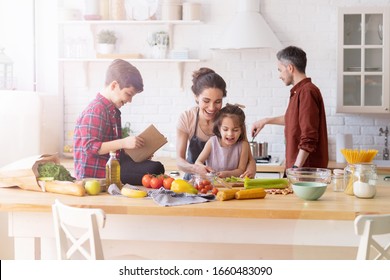 Gute Familienkochen in der Küche. Mutter und Tochter lesen Rezept für Vater und Sohn. Papa und Junge hacken grünes Pflanzenblatt für Salat. Unterkunft Erholung und Zubereitung von Speisen am Wochenende