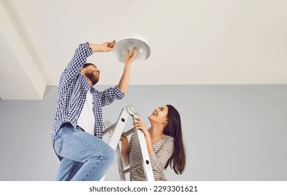 Happy family couple changing a LED lightbulb at home. Joyful young man and woman standing on a step ladder and changing an energy-saving light bulb in a white lamp on the ceiling - Powered by Shutterstock