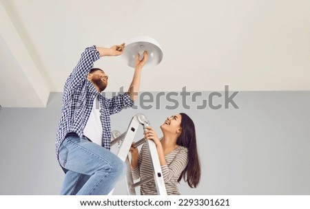 Happy family couple changing a LED lightbulb at home. Joyful young man and woman standing on a step ladder and changing an energy-saving light bulb in a white lamp on the ceiling
