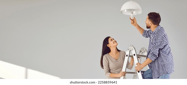 Happy family couple changing light bulb at home. Young man and woman standing on ladder and changing modern energy-saving lightbulb in ceiling lamp. Banner, header, grey copy space wall background