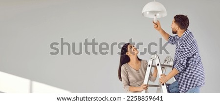 Happy family couple changing light bulb at home. Young man and woman standing on ladder and changing modern energy-saving lightbulb in ceiling lamp. Banner, header, grey copy space wall background