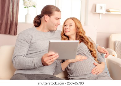 Happy family couple and digital technology. Young pregnant caucasian woman with her husband holding tablet pc on sofa at home.