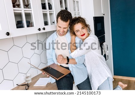 Happy family couple are standing at the kitchen at home, doing online shopping or chatting with friends. Joyful stylish husband and wife using laptop, watching funny videos or photos, smiling.