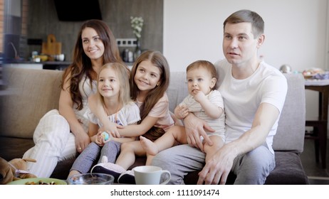 Happy Family Couple With Three Little Daughters Are Sitting On A Couch In A Living Room And Watching Tv In Daytime, Laughing And Having Fun Together