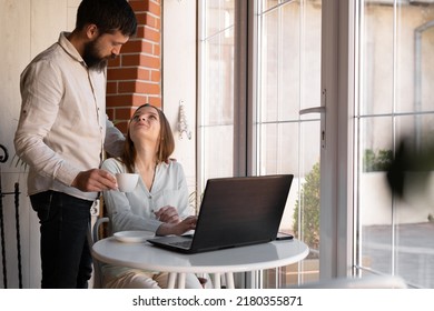 Happy Family Couple Using Laptop While Having Coffee In Modern Kitchen At Home. Young Man And Woman Hugging, Husband Give Coffee For Wife Who Work Remote