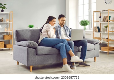 Happy family couple using laptop computer sitting on couch at home. Smiling young husband and wife looking at laptop screen while having video call, shopping online, browsing web, watching movie - Powered by Shutterstock