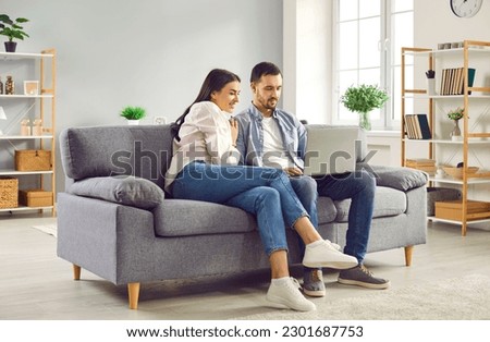 Happy family couple using laptop computer sitting on couch at home. Smiling young husband and wife looking at laptop screen while having video call, shopping online, browsing web, watching movie