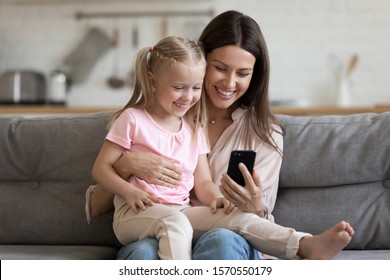 Happy Family Cute Small Kid Child Daughter Sit On Parent Mom Lap On Sofa Laugh Having Fun Hold Using Smart Phone Watch Funny Social Media Video App Take Selfie Bonding Look At Mobile Screen At Home