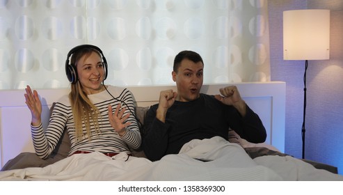 Happy Family Dancing And Listening To Music With Headphones In Bed. A Man And A Woman Are Watching TV And Having Fun.