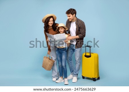 Happy family with daughter looking at map, planning travel, standing with suitcase on blue studio background, ready for summer vacation