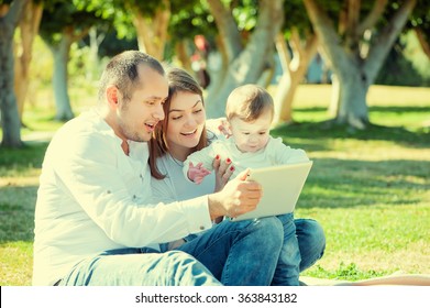 Happy Family And Digital Technology. Young Parents And Little Kid Using Table Computer While Sitting On Grass In The Park.