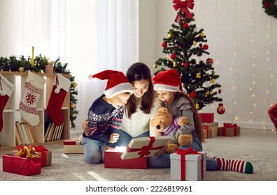 Happy family discovers wonderful surprise while opening Christmas presents. Mother and two children in red Santa hats looking inside gift box sitting together on floor in beautiful festive living room - Powered by Shutterstock