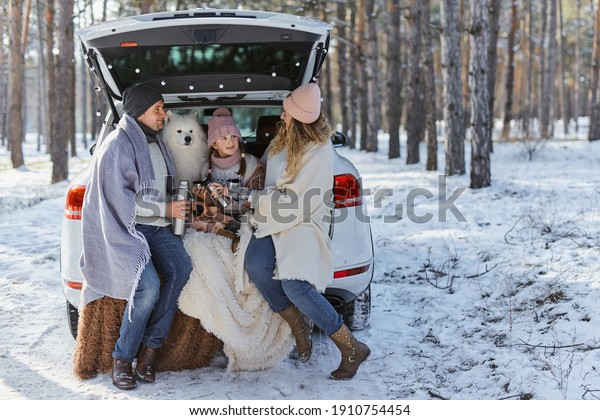 Happy\
family with dog on vacation during the winter holidays near road.\
Dressed in warm clothes sitting on the trunk of a car and drinking\
tea from a thermos. space for text. Winter\
vacation