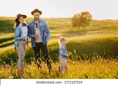 Happy Family Dressed In Casual, Enjoying Holidays In Nature During Sunset With Satisfied Smiles. Parents Wearing Hats With A Brim And Holding Glasses With White Wine Against Hills On Background.