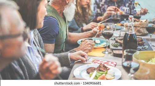 Happy family eating and drinking wine at barbecue dinner outdoor - Multiracial mature and young people having fun at bbq sunday meal - Food and summer lifestyle concept - Focus on hipster man hand