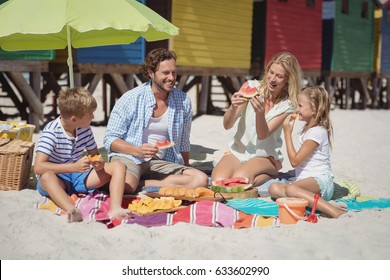 Happy family eating watermelon while sitting together on blanket at beach during sunny day - Powered by Shutterstock