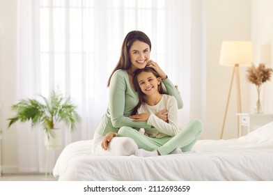 Happy family enjoying lazy morning at home. Portrait of smiling mother and child in bedroom. Beautiful mum and daughter in comfortable white and pastel green lounge homewear cuddling on cosy, warm bed