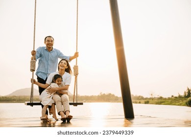 A happy family enjoying a sunny day at the park playground, with the father sitting and pushing the swing while the mother and daughter play together, Happy Family Day - Powered by Shutterstock