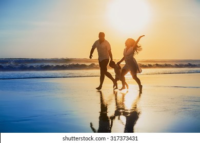 Happy family - father, mother, baby son hold hands and run with fun along edge of sunset sea on black sand beach. Active parents and people outdoor activity on tropical summer vacations with children