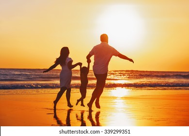 Happy Family - Father, Mother, Baby Son Hold Hands And Run With Fun Along Edge Of Sunset Sea On Black Sand Beach. Active Parents And People Outdoor Activity On Tropical Summer Vacations With Children.