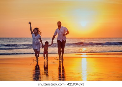 Happy family - father, mother, baby son hold hands and run with fun along sunset sea surf on black sand beach. Active parents and people outdoor activity on tropical summer vacations with children.