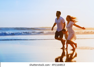 Happy family - father, mother, baby son hold hands and run together with fun along sunset sea surf on black sand beach. Travel, active lifestyle, parents with children on tropical summer vacations.
