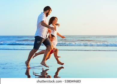 Happy Family - Father, Mother, Baby Son Hold Hands, Run Together With Splashes By Water Pool Along Sunset Sea Surf On Black Sand Beach. Travel, Active Lifestyle, Parents With Child On Summer Vacation.