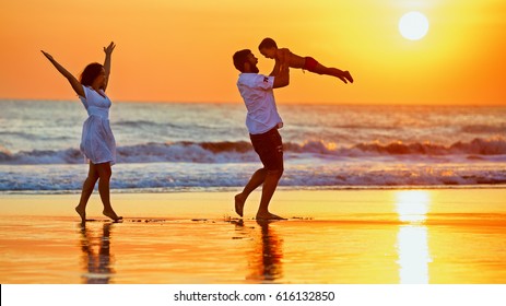 Happy Family - Father, Mother, Baby Son Walk With Fun Along Edge Of Sunset Sea Surf On Black Sand Beach. Active Parents And People Outdoor Activity On Summer Vacations With Children On Bali Island