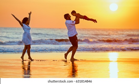 Happy Family - Father, Mother, Baby Son Walk With Fun Along Edge Of Sunset Sea Surf On Black Sand Beach. Active Parents And People Outdoor Activity On Summer Vacations With Children On Bali Island