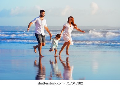 Happy family - father, mother, baby son have fun together, child run with splashes by water pool along sunset sea surf on black sand beach. Travel lifestyle, parents with kids on summer vacation.