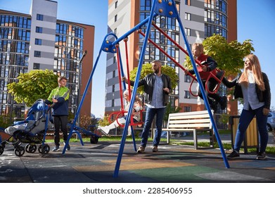 Happy family - father, mother and children having fun together at modern courtyard of city residential high-rise buildings. Parents swinging daughter and son, while other child standing with pram.