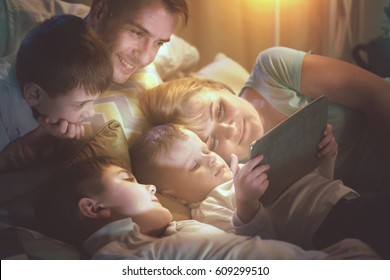 Happy Family- Father, Mother And Three Kids Lying Down On The Sofa And Using Tablet Pc At Night. Big Family Together Watching Movie On Tablet Computer In Dark Room. 