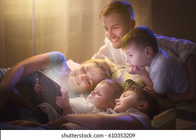 Happy Family- Father, Mother And Three Kids Lying Down On The Sofa And Using Tablet Pc At Night. Big Family Together Watching Movie On Tablet Computer In Dark Room. Togetherness 