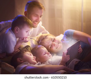 Happy Family- Father, Mother And Three Kids Lying Down On The Sofa And Using Tablet Pc At Night. Big Family Together Watching Movie On Tablet Computer In Dark Room. Bedtime