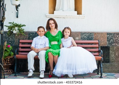 Happy Family At First Holy Communion Sitting On Bench Background Church. Mother With Sun And Daughter