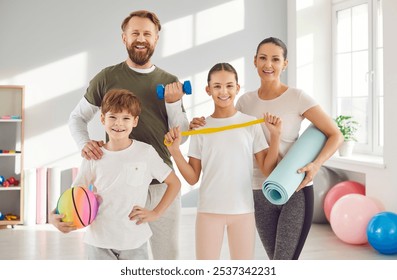 Happy family of four enjoying a fitness workout together. Cheerful, confident mum, dad and children standing in the gym, holding sports equipment, looking at the camera and smiling - Powered by Shutterstock