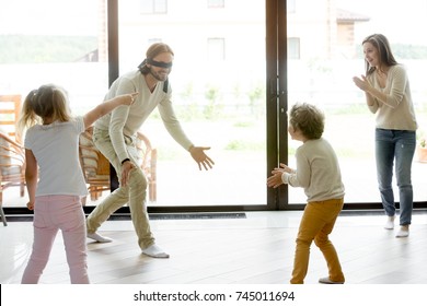 Happy family of four playing funny hide and seek game at home, blindfolded father trying to catch mother or children, parents and kids having fun laughing spending free time together in living room
