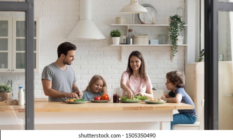 Happy Family Of Four Preparing Healthy Food Salad Together At Kitchen. Smiling Young Parents Chopping Vegetables While Little Preschool Children Waiting For Food, Communicating, Enjoying Weekend Time.