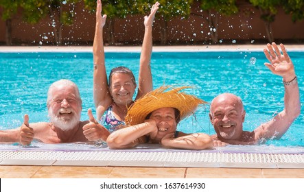 Happy Family Of Four Senior People Floating In Outdoor Swimming Pool Raising Splashes Of Water. They Smile Relaxed On Vacation Under The Bright Sun