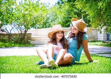 Happy Family In The Garden, Beautiful Mother With Her Little Cute Daughter Sitting On Fresh Green Grass On Backyard, With Pleasure Spending Time Together