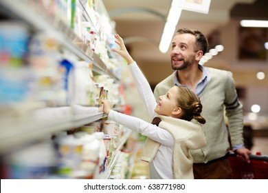 Happy family grocery shopping in supermarket: smiling man with daughter choosing dairy products from fridge in milk aisle