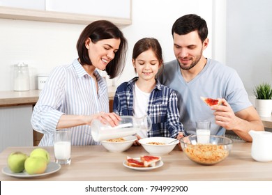 Happy Family Have Healthy Breakfast Together. Smiling Mother Pours Milk In Bowl With Cornflakes, Eat Apples, Snacks And Drink Milk, Sit Together At Kitchen. Healthy Food, Family And Eating Concept.