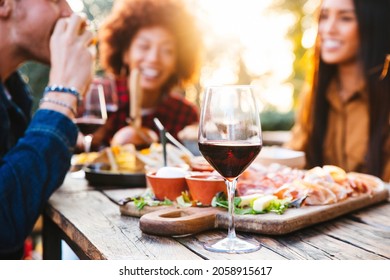 Happy Family Having Barbecue Party In Backyard - Young Friends Celebrating At Dinner Drinking Red Wine At Sunset - Focus On Wine Glass