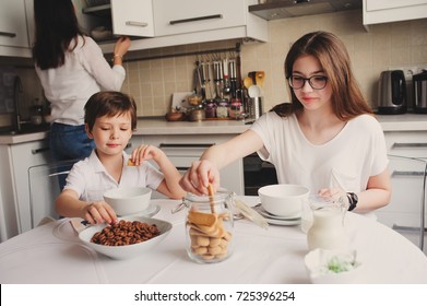 Happy Family Having Breakfast At Home. Mother With Two Kids Eating In Modern White Kitchen