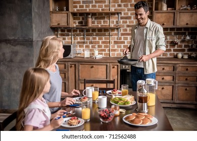 Happy Family Having Breakfast In Kitchen. Mom And Daughter Are Sitting At The Table While Dad Is Making Pancakes