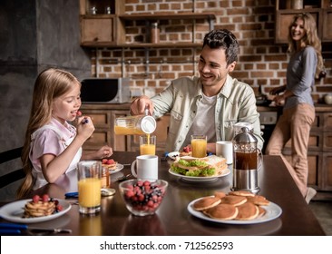 Happy Family Having Breakfast In Kitchen. Dad And Daughter Are Sitting At The Table While Mom Is Making Pancakes