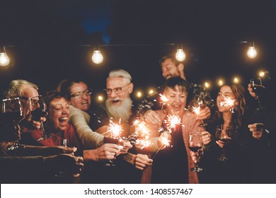 Happy family having at dinner party outdoor - Group of multiracial older and young people celebrating together drinking wine holding fireworks sparklers - Concept of youth and elderly parenthood