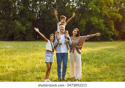 Happy family having fun in a fresh green summer park. Full body length portrait of cheerful, joyful mother, father and children standing together on a grassy lawn, waving hello and smiling  - Powered by Shutterstock