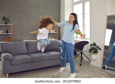 Happy Family Having Fun At Home. Mother And Little Child Playing In Living Room With Sound Proof Walls Without Disturbing Neighbors. Smiling Mommy Holding Daughter's Hand While She Is Jumping Off Sofa